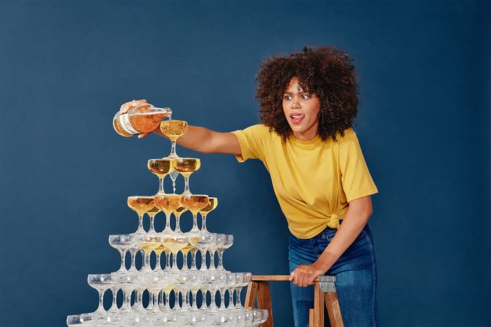 Woman in yellow pours champagne into a tower of glasses against a blue background, creating a celebratory and dynamic visual.