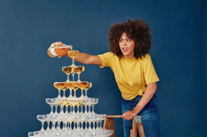 Woman in yellow pours champagne into a tower of glasses against a blue background, creating a celebratory and dynamic visual.
