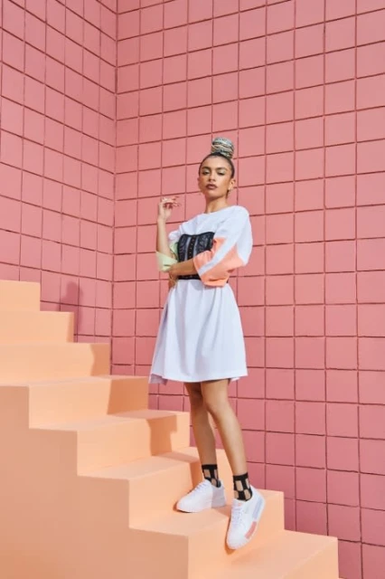 Woman in a white T-shirt dress and sneakers poses on peach-colored stairs against a tiled pink wall, looking confidently at the camera.