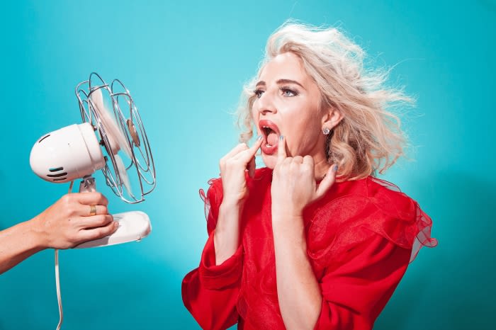 Woman in red opens mouth in front of electric fan held by arm on blue background to illustrate staying cool in the summer.