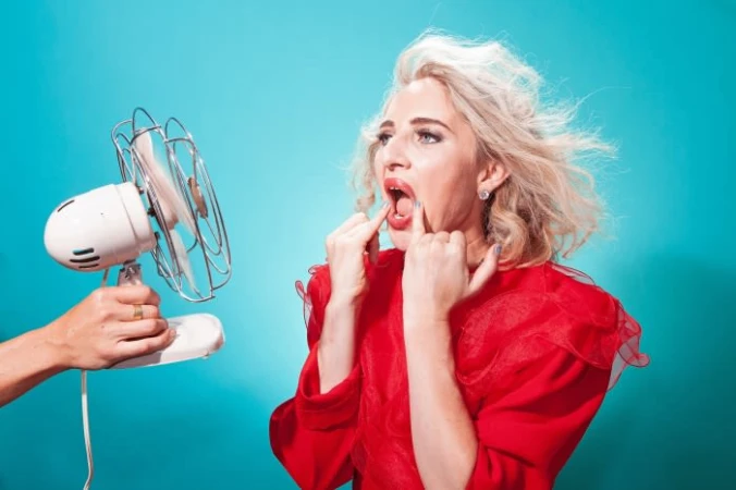 Woman in red opens mouth in front of electric fan held by arm on blue background to illustrate staying cool in the summer.