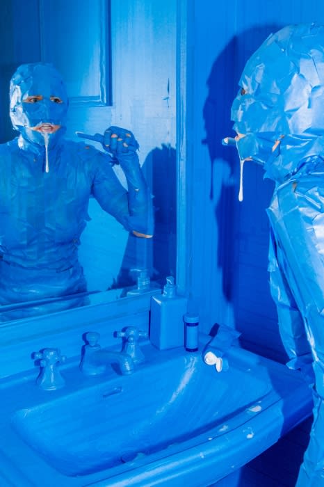 A person in a blue bodysuit stands in a blue bathroom, looking at their reflection in the mirror while brushing their teeth.