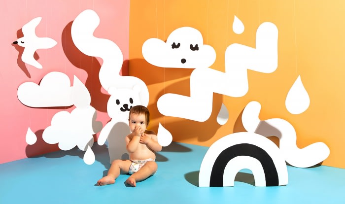 Infant sits amidst playful cloud and rainbow cutouts in a colorful studio setting, creating a whimsical and imaginative scene.