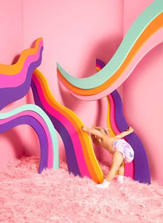 Blonde toddler in diaper stands in a pink studio against pink shag carpet with colorful archway art installations behind her.