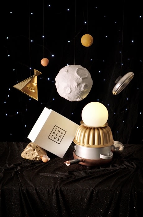 An imaginative still-life composition featuring a robot constructed from kitchenware, positioned on a shimmering black fabric against a starlit backdrop.