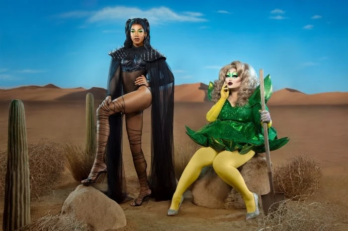 Two drag queens pose in a desert landscape. One stands in a black bodysuit with long cape and thigh-high boots. The other sits in a green dress.