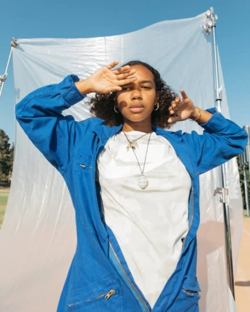 A woman poses outdoors in a blue jumpsuit, shielding her eyes from the sun. She wears jewelry with a translucent backdrop and blue sky.