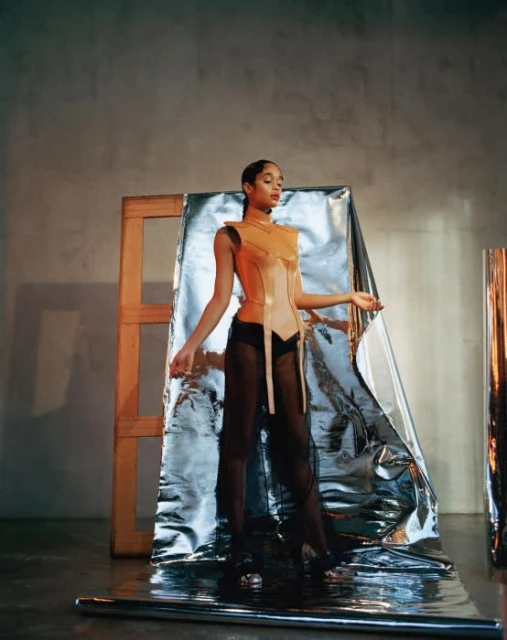 Woman poses in a sculptural orange corset top and flowing black pants, with reflective material and a wood frame in the background.
