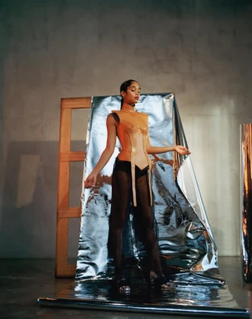 Woman poses in a sculptural orange corset top and flowing black pants, with reflective material and a wood frame in the background.