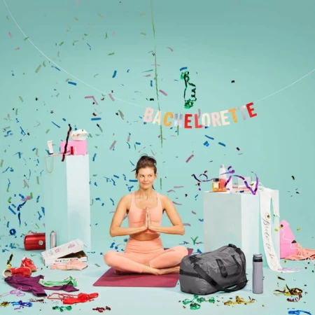 Woman in a yoga pose seated on a mat with a duffel bag and party decor on a studio set against a light blue background.