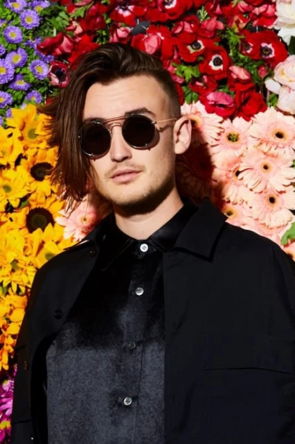 Portrait of a stylish adult male with sunglasses in front of a flower wall, wearing a black jacket and patterned shirt. Cool, fashionable.