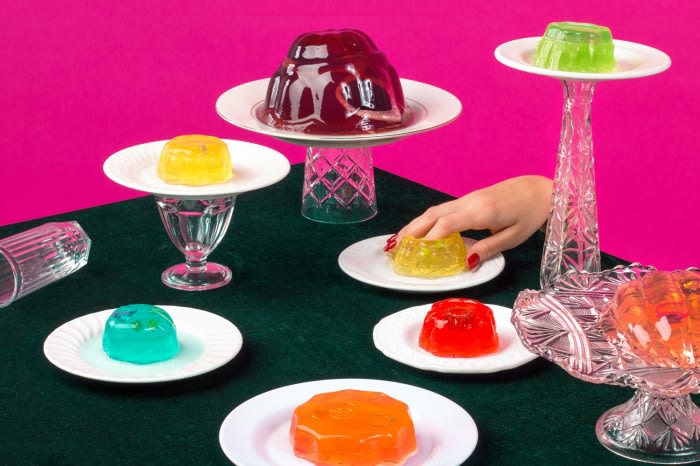 Colorful assortment of gelatin desserts on plates, set on a green surface against a vibrant pink background, with a hand reaching for one.
