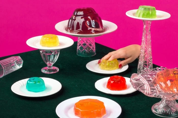 Colorful assortment of gelatin desserts on plates, set on a green surface against a vibrant pink background, with a hand reaching for one.