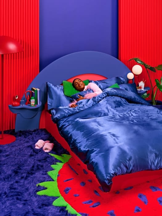 Person sleeping in a blue bed in a bright red and blue room with books and decor, illuminated by a lamp. 