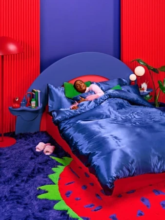 Person sleeping in a blue bed in a bright red and blue room with books and decor, illuminated by a lamp. 