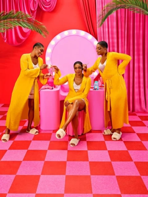 Three dark-skinned women pose in a vibrant photo studio with red and pink colors, wearing yellow robes and white slippers.