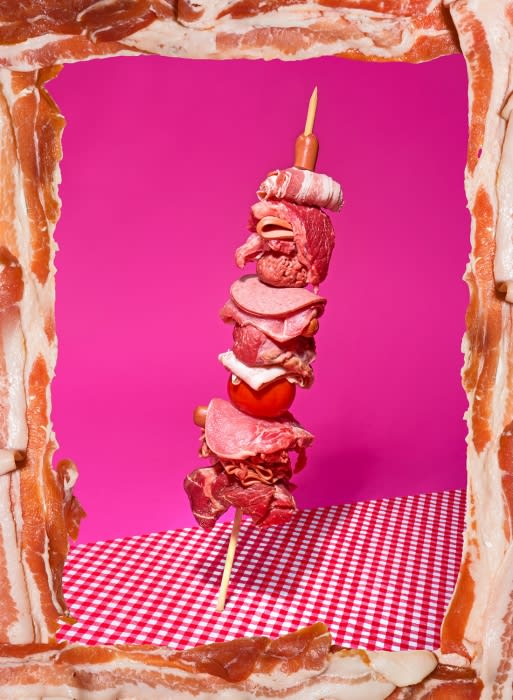 Creative food photography of meat and tomato skewer on a red gingham tablecloth, framed by bacon strips against a pink background.