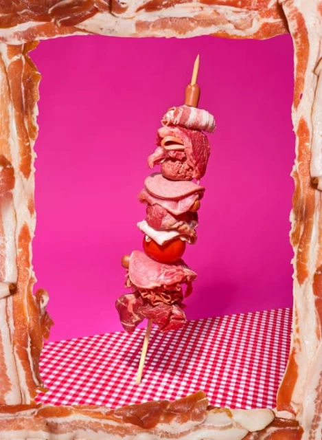 Creative food photography of meat and tomato skewer on a red gingham tablecloth, framed by bacon strips against a pink background.