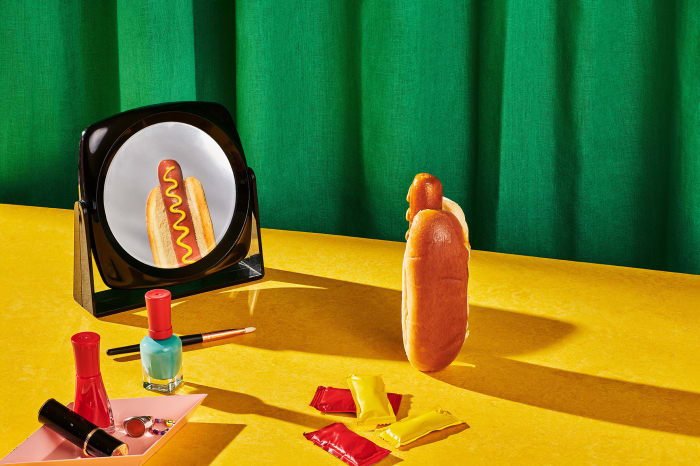 Whimsical still life featuring a hot dog beside a mirror reflecting a hot dog, alongside makeup and condiments on a yellow surface.