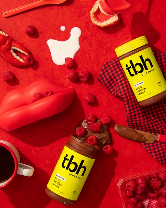 Two jars of hazelnut cocoa spread surrounded by raspberries and red kitchen objects on a red background.