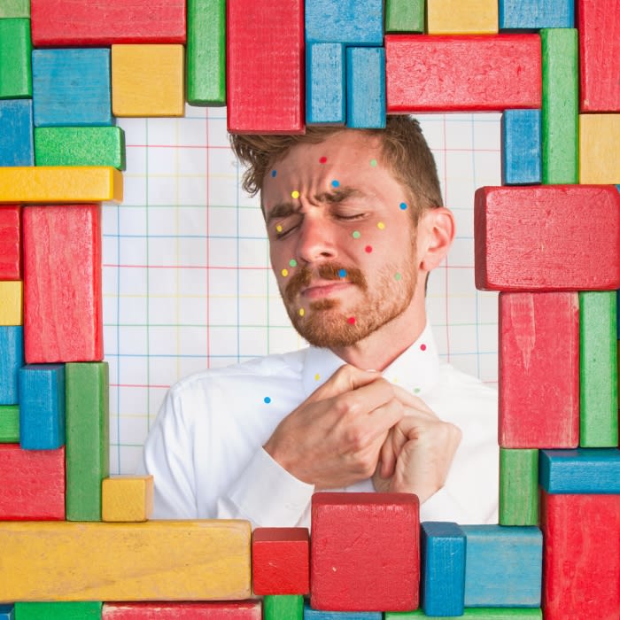 Man with colorful dots on his face framed by colorful building blocks against a grid background, eyes closed and hands clasped.