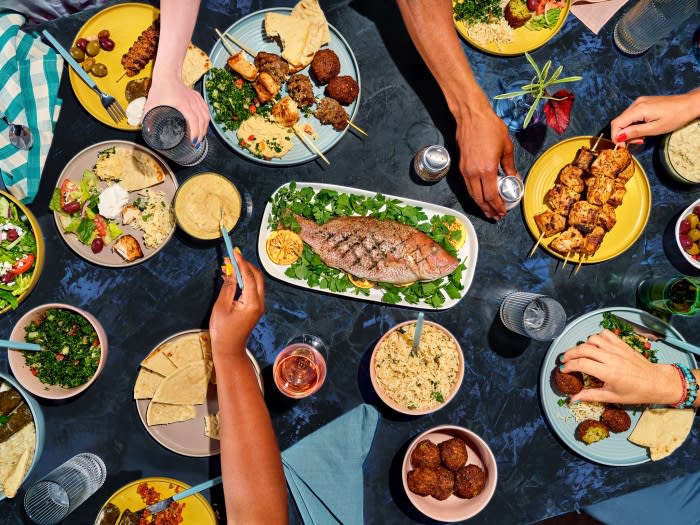 Overhead shot of a group gathering around a table laden with Middle Eastern cuisine including grilled fish, kebabs, and fresh salads.