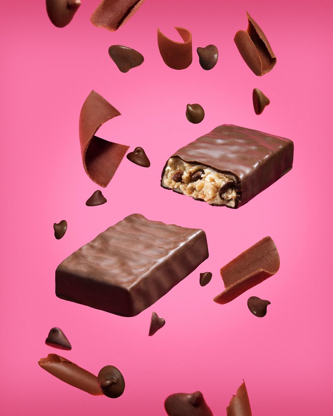 Two chocolate-covered protein bars float with chocolate chips and shavings against a bright pink background in a studio shot.