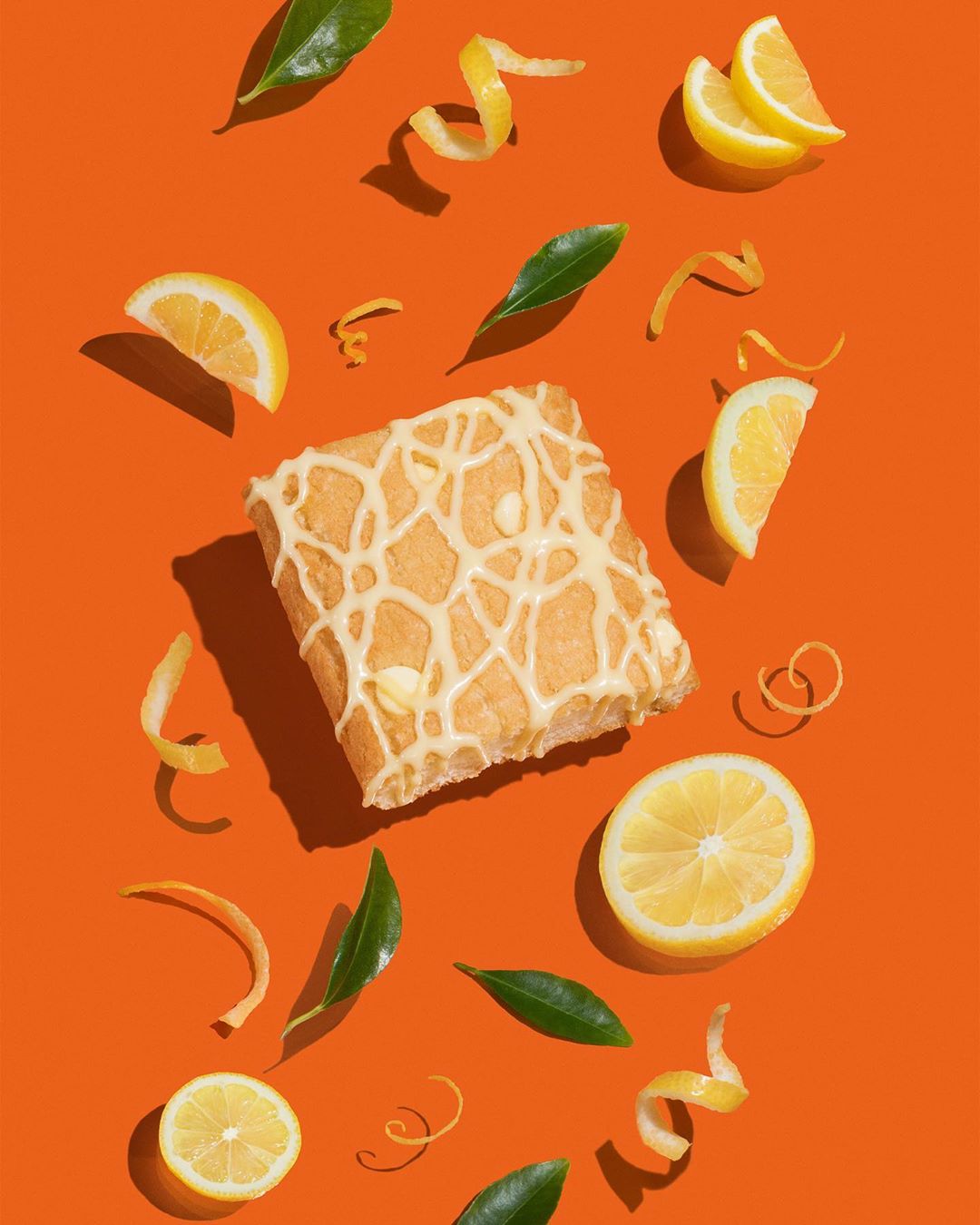 Overhead shot of a glazed pastry with lemons, peels, and green leaves on a vibrant orange backdrop, styled for food photography.