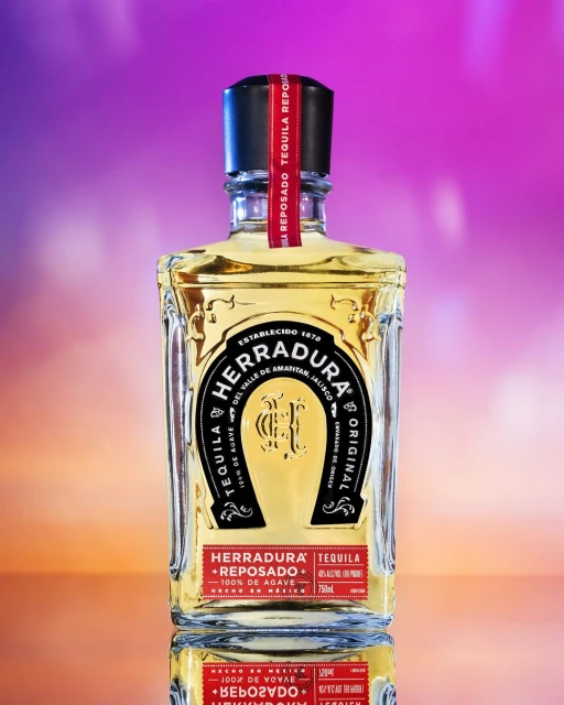 Bottle of Herradura Reposado tequila on a reflective surface, with a gradient purple and orange background. Liquor bottle is the focus.