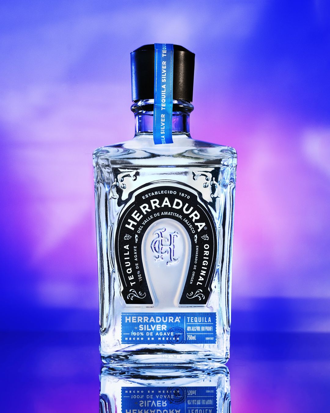 Crystal Herradura Silver Tequila bottle with blue label on a reflective surface and a soft blue and purple background. 