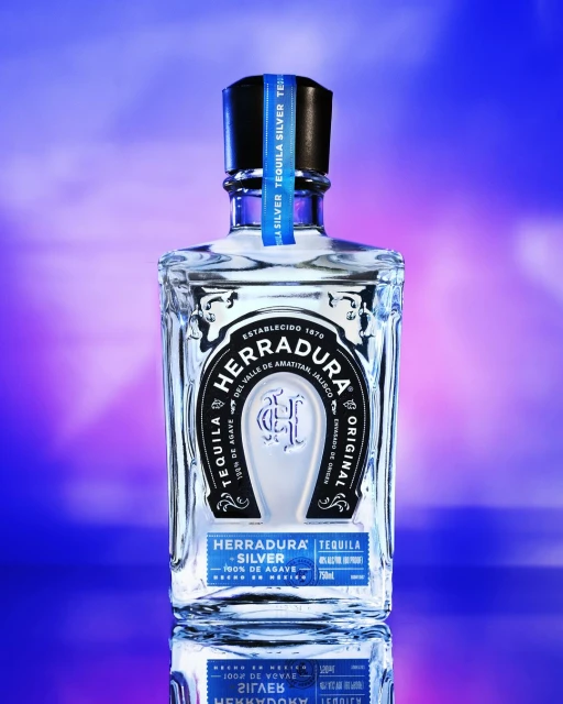 Crystal Herradura Silver Tequila bottle with blue label on a reflective surface and a soft blue and purple background. 
