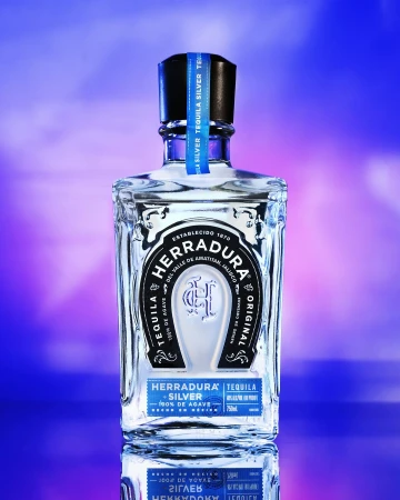 Crystal Herradura Silver Tequila bottle with blue label on a reflective surface and a soft blue and purple background. 