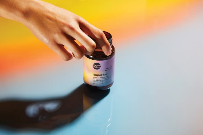 A hand reaches for a bottle of SuperYou dietary supplements, product shot on a split yellow and blue background with a soft shadow.