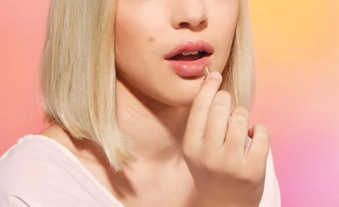 Woman with blonde hair putting capsule medication into her mouth against a blended pink and yellow gradient background in a beauty shot.