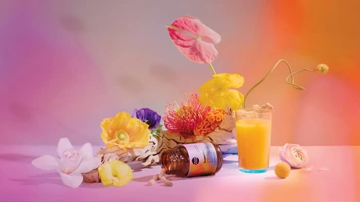Botanical still life featuring orange juice, vitamin bottle, capsules, and colorful assorted flowers against a vibrant gradient backdrop.