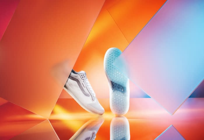 Studio shot of a white sneaker with grey details shown with its sole and placed against an orange and blue geometric background.