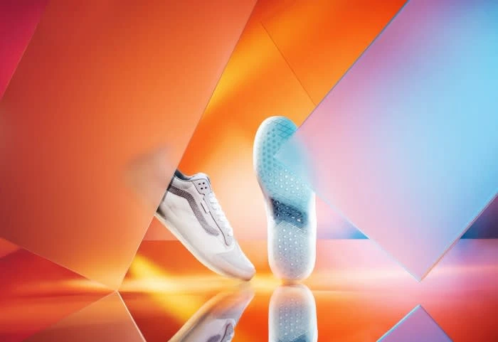Studio shot of a white sneaker with grey details shown with its sole and placed against an orange and blue geometric background.