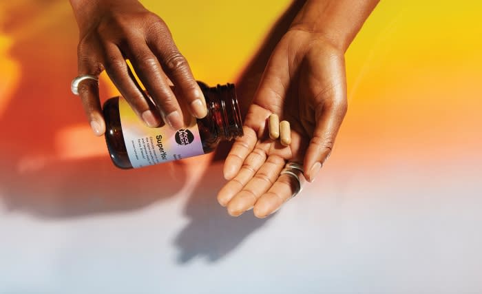 Dark-skinned hands pour tan capsules from a brown supplement bottle against a yellow-to-white gradient background with shadows.