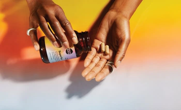 Dark-skinned hands pour tan capsules from a brown supplement bottle against a yellow-to-white gradient background with shadows.
