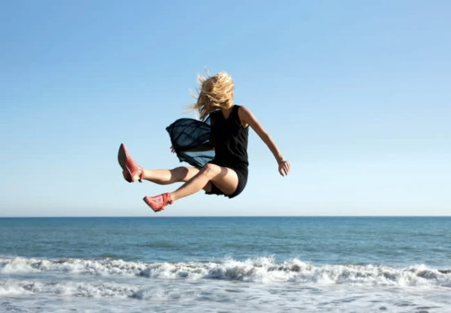 Blonde woman in black dress and red shoes mid-air jumping over ocean waves under clear blue sky