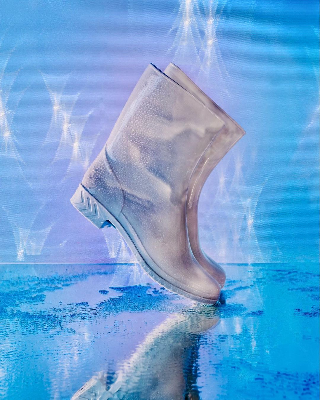 Rain boots on a reflective wet surface against a light blue background with water-like light effects overhead, studio product shot.