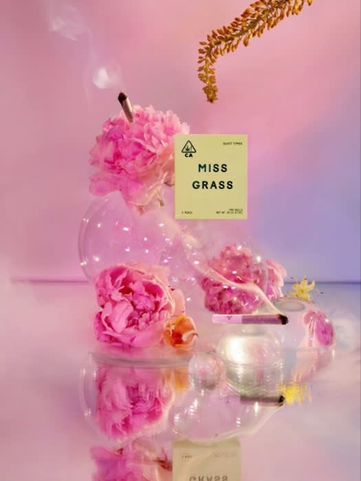 Artistic cannabis product shot with pink peonies, laboratory glassware, and joint with rising smoke, soft pink and blue gradient background.