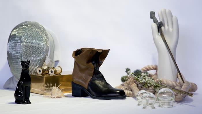 Still life composition featuring a heeled boot, fencing mask, a black cat figurine, and other objects against a white background.