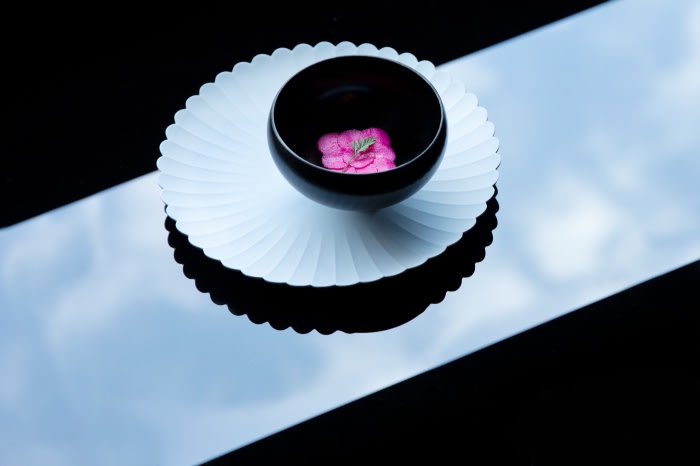 Still life of a black bowl with pink flowers on a white plate, set on a glossy black surface, reflecting sky and clouds.