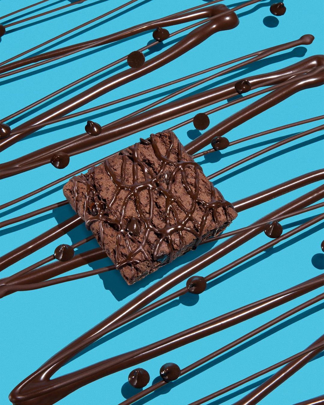 Square brownie topped with chocolate drizzle on a bright blue surface with chocolate drizzle and drops, close-up food photography.