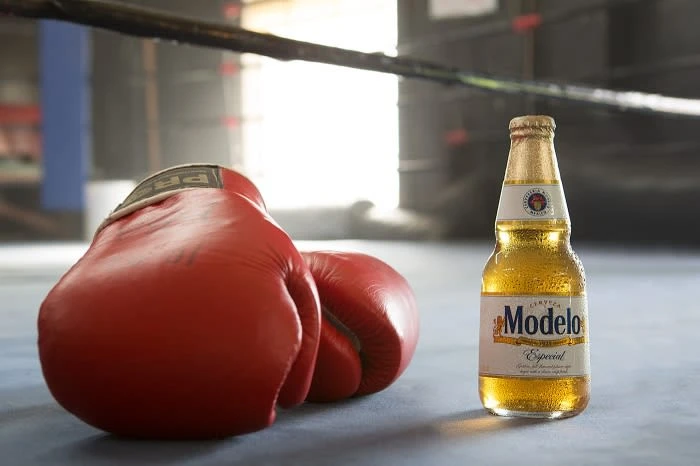 Bottle of Modelo Especial beer stands near a red boxing glove on a light grey surface in a boxing ring setting with bright lighting.