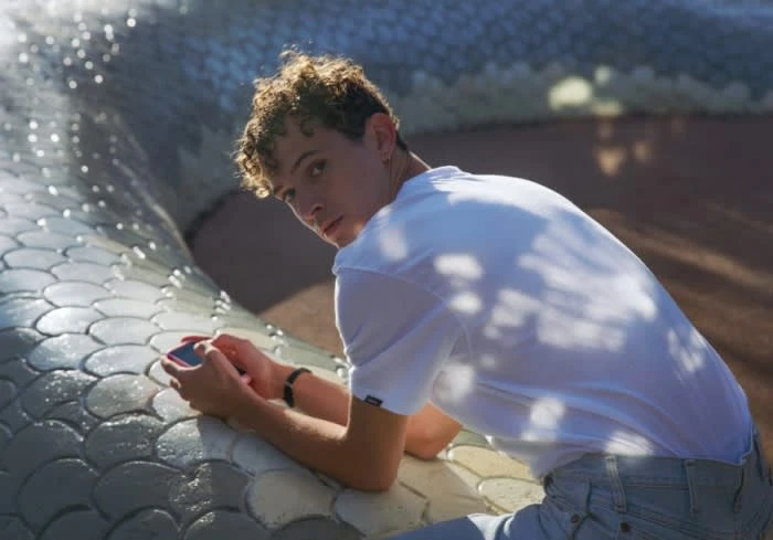 Young man with curly hair leans against a silver, scaled structure, holding a mobile phone, in bright daylight.