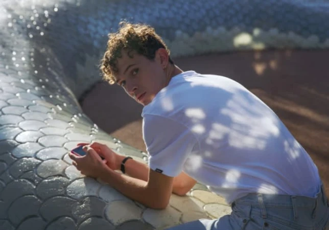 Young man with curly hair leans against a silver, scaled structure, holding a mobile phone, in bright daylight.
