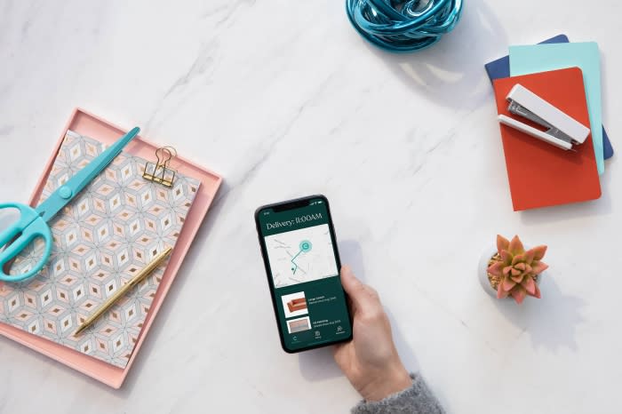 Overhead shot of hand holding smartphone with delivery app, surrounded by scissors, pen, notebooks, stapler, succulent, and yarn on marble.