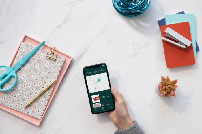 Overhead shot of hand holding smartphone with delivery app, surrounded by scissors, pen, notebooks, stapler, succulent, and yarn on marble.